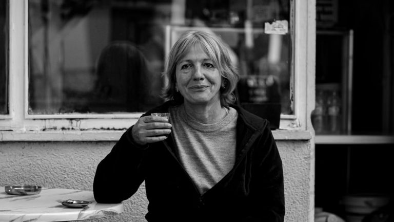Black and white portrait of a senior woman with a coffee cup, sitting outside a café.